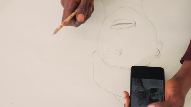Artist finalizing portrait sketch with black painting reference on phone and pencil on paper. Wide composition shows hand refining eyes and mouth, subtle shading, intentional strokes and quiet studio