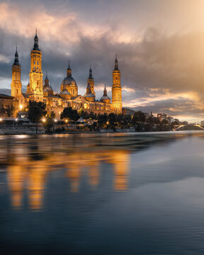 View of the Cathedral-Basilica of Our Lady of the Pillar reflecting in the Ebro River under a dramatic sunset sky in Zaragoza, Aragon, Spain.