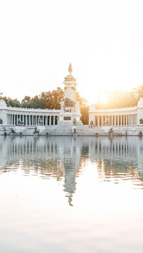 View of the Monument to Alfonso XII with its white colonnade reflected in the calm waters of the Retiro Park lake during golden hour in Madrid, Community of Madrid, Spain.