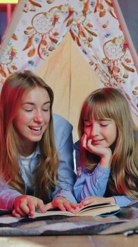 Happiness concept where lovely cheerful satisfied eldest and younger sisters lying on coverlets in makeshift wigwam in playroom with night lightings and reading interesting book