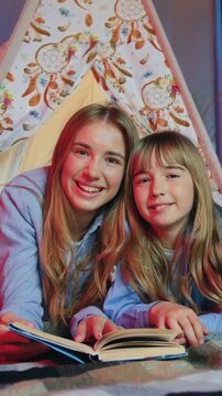 Sisters leisure concept where attractive friendly joyful loving eldest and younger girls lying on coverlets in makeshift wigwam and enjoying of reading interesting book,posing on camera