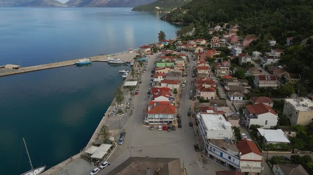 Coastal town with a harbor, lined with houses and trees, overlooking a serene blue sea and distant mountains.