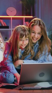 Leisure concept where beautiful satisfied friendly blond eldest and younger sisters having fun together in night room interior on the floor on bedcovers and applying computer to revision joint photos