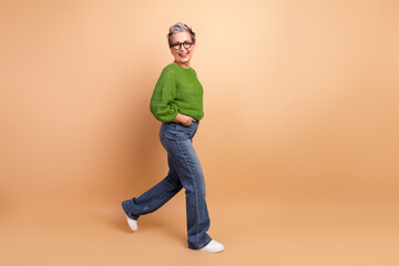 Mature stylish lady in green sweater and jeans walks in studio against beige background smiling...