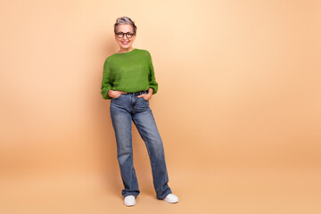 Confident mature lady in green sweater and jeans stands against beige background smiling warmly for...