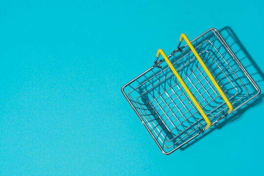 Flat lay of empty metal shopping basket with yellow handles on bright turquoise blue background. Concepts of shopping and sale season.