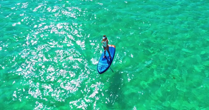 Aerial view of woman in white swimsuite standing on sup board with paddle on crystal turquoise water with coral reef on the tropical island in Indian ocean. Active water sport. Mauritius island