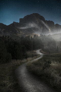 View of a winding gravel path leading towards a massive mountain peak under a starry night sky in South Tyrol, Trentino-South Tyrol, Italy.
