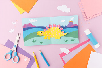 Paper cut dinosaur in landscape with mountains and volcano on folded card with scissors, glue stick, pencils and colored paper on pink background. Flat lay with copy space. Kids craft concept.  © Elena