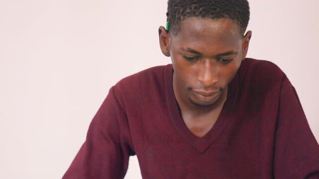 Black male artist planning painting composition, contemplative pose with phone reference and pencil, maroon sweater at quiet table, calm indoor studio atmosphere, thoughtful expression showing early