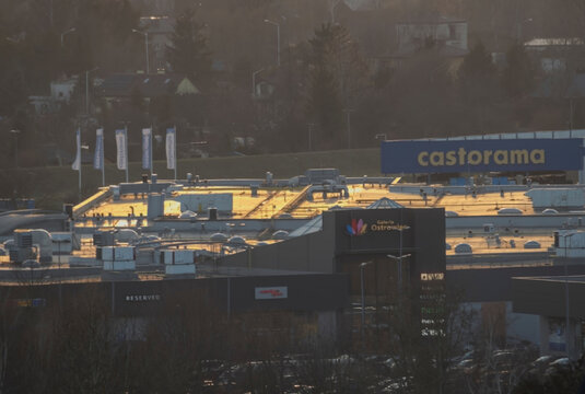 Polska, Ostrowiec Świętokrzyski, 18 marca 2026, 17:33. Castorama i Galeria Ostrowiec w blasku zachodzącego słońca. Widok na centrum handlowe o zachodzie słońca. Konstrukcje dachowe i szyldy.