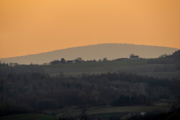 Wieczór zapada nad Górami Świętokrzyskimi w ostatnich dniach zimy w Polsce, rzucając ciepły, pomarańczowy odcień. Krajobraz charakteryzuje się łagodnymi wzgórzami i drzewami, oraz domami © Grzegorz