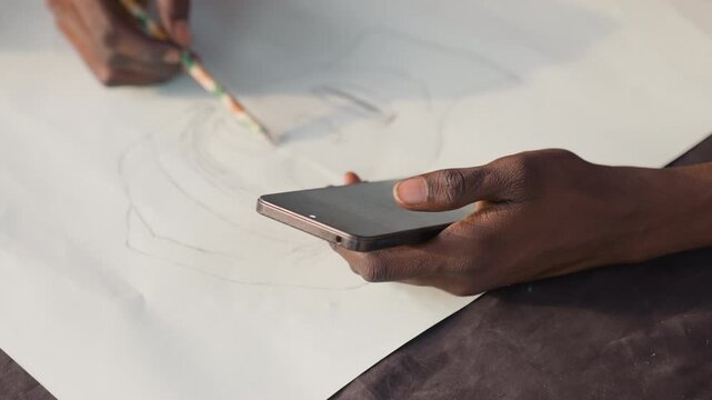 Hands sketching on large paper black painting study with phone reference brush tracing charcoal lines closeup of fingertips portrait outline emerging artist planning pattern and composition focused