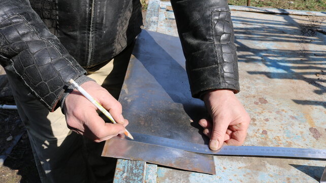 Marking a small sheet of iron on an outdoor workbench using a metallic ruler and a pencil, taking measurements of a part on a plane, and planning a metal object