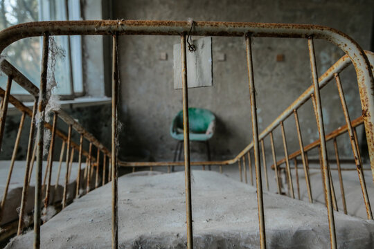 View of a rusty metal bed frame with a hanging tag in an abandoned hospital room with a dusty mattress and a green chair Pryp'yat', Kyiv Oblast, Ukraine.