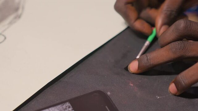 Closeup hands etching black painting mat, artist using green tool to scrape pigment from textured surface next to smartphone, red paint specks, precise motion, focused tactile studio atmosphere