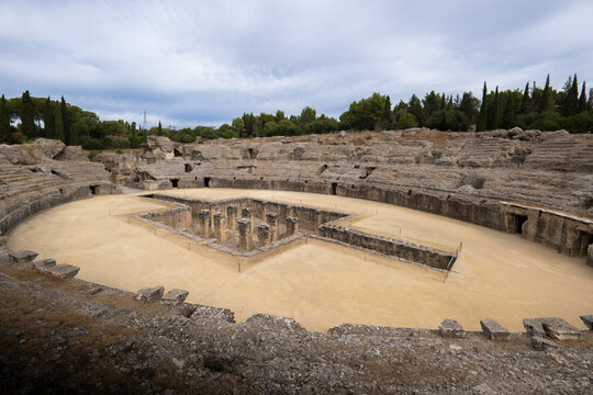 Santiponce, Spain - 13 August 2024: View of the Roman amphitheater at the Conjunto Arqueologico de Italica with ancient stone tiers and central arena pits under a cloudy sky.