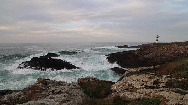 View of Faro Illa Pancha lighthouse standing on a rugged rocky cliff with waves crashing against the shore under an overcast sky in Isla Pancha, Lugo, Spain.