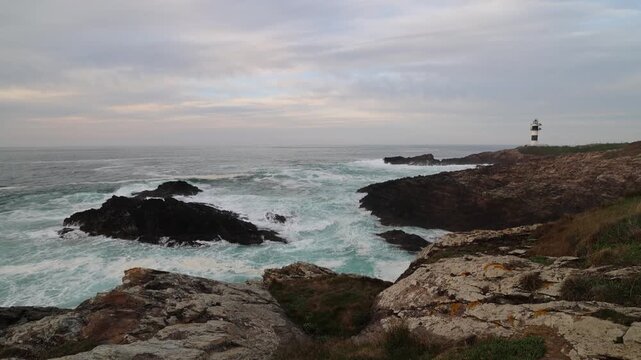 View of Faro Illa Pancha lighthouse on a rocky cliff with waves crashing against the shore under a cloudy sky Isla Pancha, Lugo, Spain.