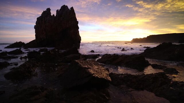 View of the dramatic rock formations at Playa de Portizuelo with waves crashing against the shore during a colorful sunset in Luarca, Asturias, Spain.