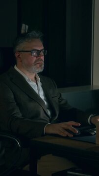 Medium vertical shot of frustrated Caucasian businessman working on documents on computer in dark office in middle of night, talking off glasses and rubbing dry eyes, suffering from burnout