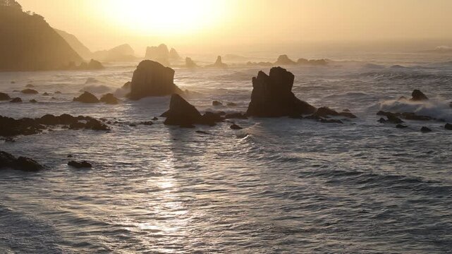 View of Silence Beach with dramatic sea stacks and waves crashing under a golden sunset light in Asturias, Asturias, Spain.