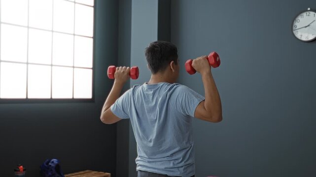 Man raises red dumbbells overhead with forearms visible by large window and wall clock in a studio building; determination.