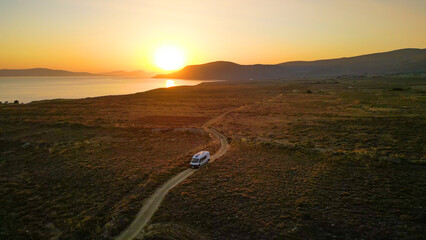 A camper van driving along a remote coastal road at sunset on Limnos Island, Greece. Aerial perspective shows golden Mediterranean light, rugged landscape and freedom of van life travel. © _jure