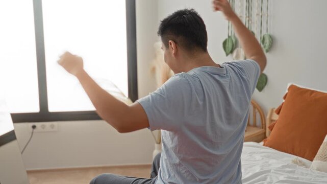 Man on bed with hands clasped behind neck stretching seated on sheets in building; calm morning routine.
