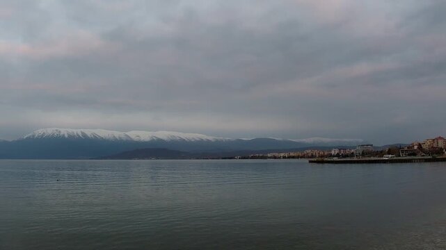 A cinematic zoom-in timelapse of the Pogradec shoreline on Lake Ohrid, Albania, featuring the city's waterfront architecture against a backdrop of snow-capped mountains.