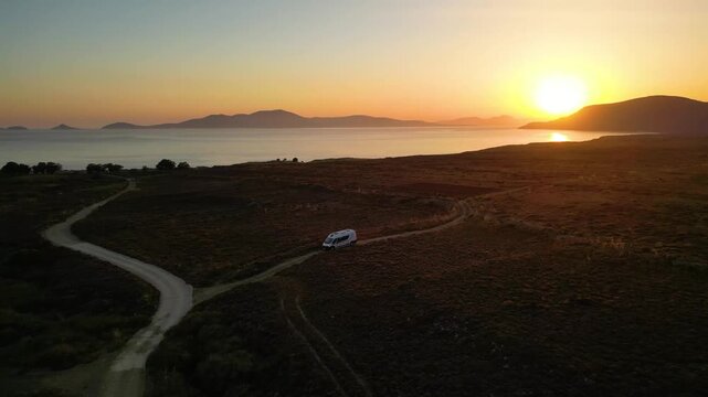 A camper van driving along a remote coastal road at sunset on Limnos Island, Greece. Aerial perspective shows golden Mediterranean light, rugged landscape and freedom of van life travel.