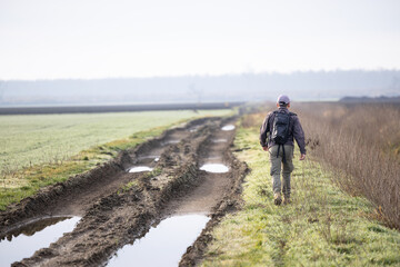 Obraz premium Hiker Walking Along Muddy Dirt Road Through Frosty Field With Backpack On Overcast Morning
