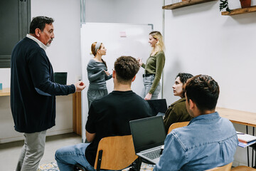 Naklejka premium hispanic senior man teacher with multicultural group of university students talking in classroom. Diverse group of people in Education concept