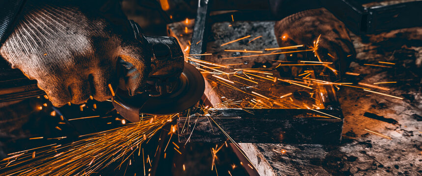 Unrecognizable worker hand in protective glove using an angle grinder tool to cut a metal piece, generating bright orange and yellow sparks during the industrial process.