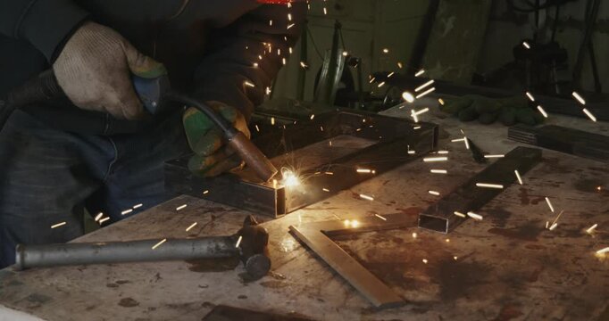 Professional welder joining metal profile pipes with an arc welding machine in a workshop, with bright sparks flying from the molten metal as he fabricates a steel frame on a workbench.
