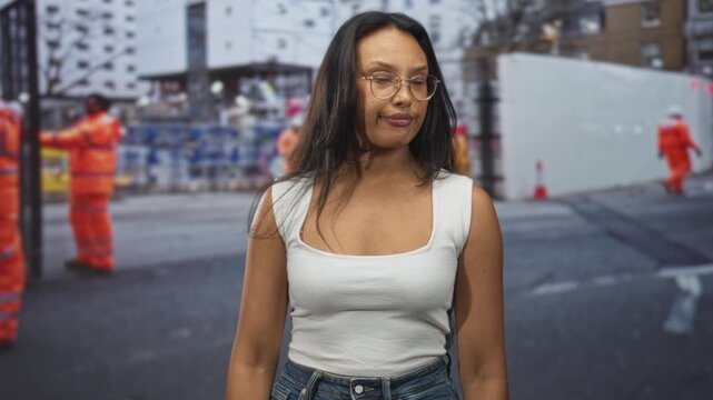Woman in white tank top wearing glasses tilts head with a smirk on a street with construction workers in view; urban attitude amused.