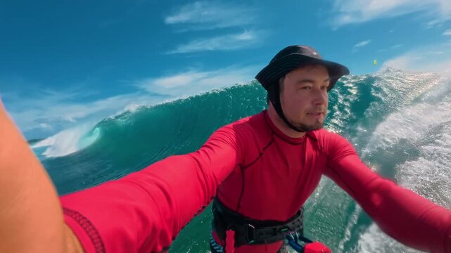 A male kitesurfer in a red sweater and sunhat films himself riding a huge ocean wave. Extreme sports