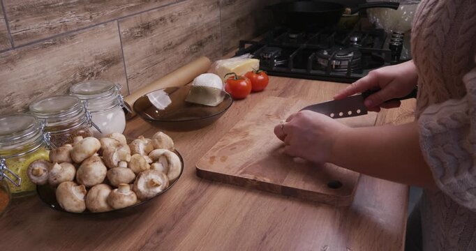 Female hands using a sharp knife to carefully chop fresh champignon mushrooms on a wooden board in a cozy home kitchen, preparing ingredients for a delicious and healthy homemade meal.