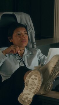 Medium vertical shot of Hispanic businesswoman studying document attentively while sitting at desk with feet up at night in dark office, then thinking while fidgeting with glasses