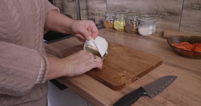 Unrecognizable woman unwrapping a wheel of fresh brie or camembert cheese on a wooden cutting board in a domestic kitchen, preparing to slice the delicious dairy product for a meal.