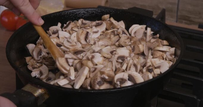 Woman's hand stirring sliced champignon mushrooms with a wooden spatula while frying them in a black cast iron skillet on a gas stove, preparing a delicious homemade vegan meal.