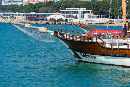 Wooden sailing ship with name "Korsar" docks in clear blue water, with modern buildings and pier surrounded by greenery in background. Gelendzhik, Russia - March 2, 2026