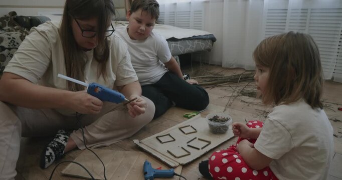 Loving family sitting on the floor making a creative art project using natural materials, with the mother using a hot glue gun to attach small twigs onto cardboard while her children watch.
