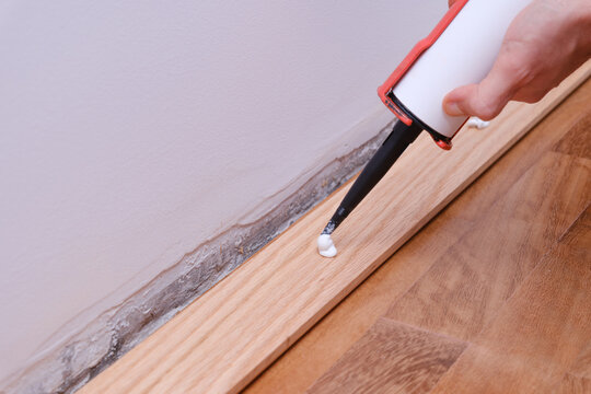 Hand applying white adhesive along a baseboard with a caulking gun