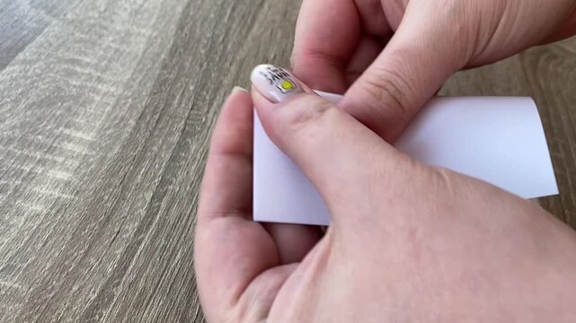 Female hands folding blank white paper card on wooden table, minimal close up composition with neat nail art, simple craft process, clean neutral styling and realistic everyday gesture