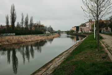 Obraz premium Wide view of river with trees and buildings in Zrenjanin, Serbia under cloudy sky. European landscape and calm city atmosphere concept.