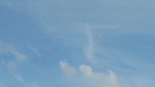 A rocket climbs high into a blue sky surrounded by soft clouds with a bright flame trail visible below. The spacecraft moves upward through the atmosphere leaving a glowing streak against the open sky