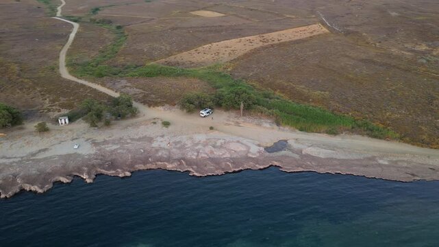 Aerial view of Paralia Skidi beach on Limnos Island, Greece, featuring a parked campervan beside a remote coastline. Clear Mediterranean water and rugged terrain highlight peaceful wild camping
