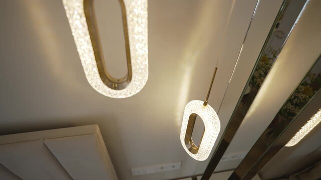A smooth low angle shot moving towards modern oval shaped crystal and gold pendant lights hanging in a luxurious bedroom. Elegant interior design and warm, ambient lighting create a cozy atmosphere