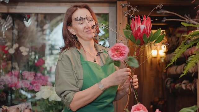 Smiling woman showcases floral arrangement. Friendly florist offers vibrant flower stems in cozy shop setting. Happy female florist presents boisterous protea bunch amid welcoming shop ambiance